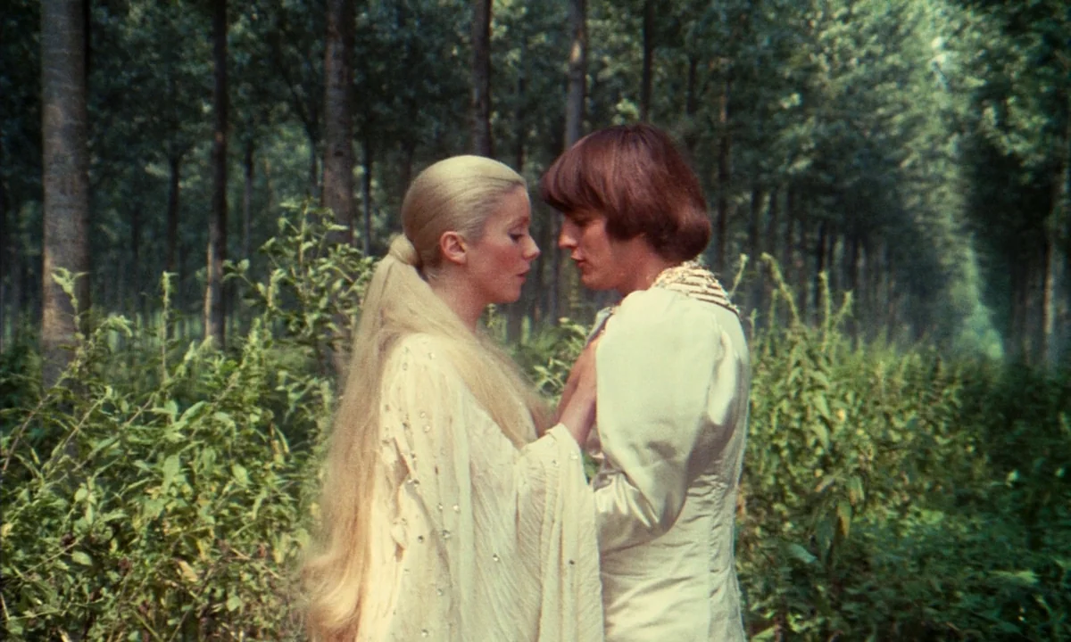 Donkey Skin (1970) movie still: medium — A dense grove of young trees with tall vertical trunks and lush foliage ground. A man and a woman in white…