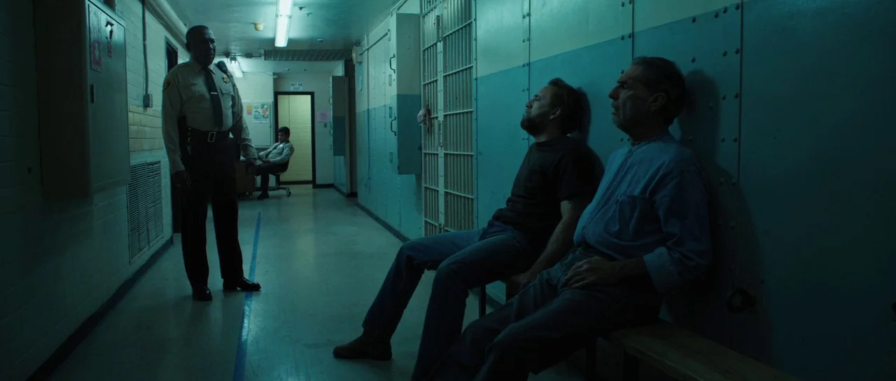 Joe (2013) movie still: fullbody — A long institutional hallway features teal walls, metal barred doors, and fluorescent lighting. Two men sit on a…