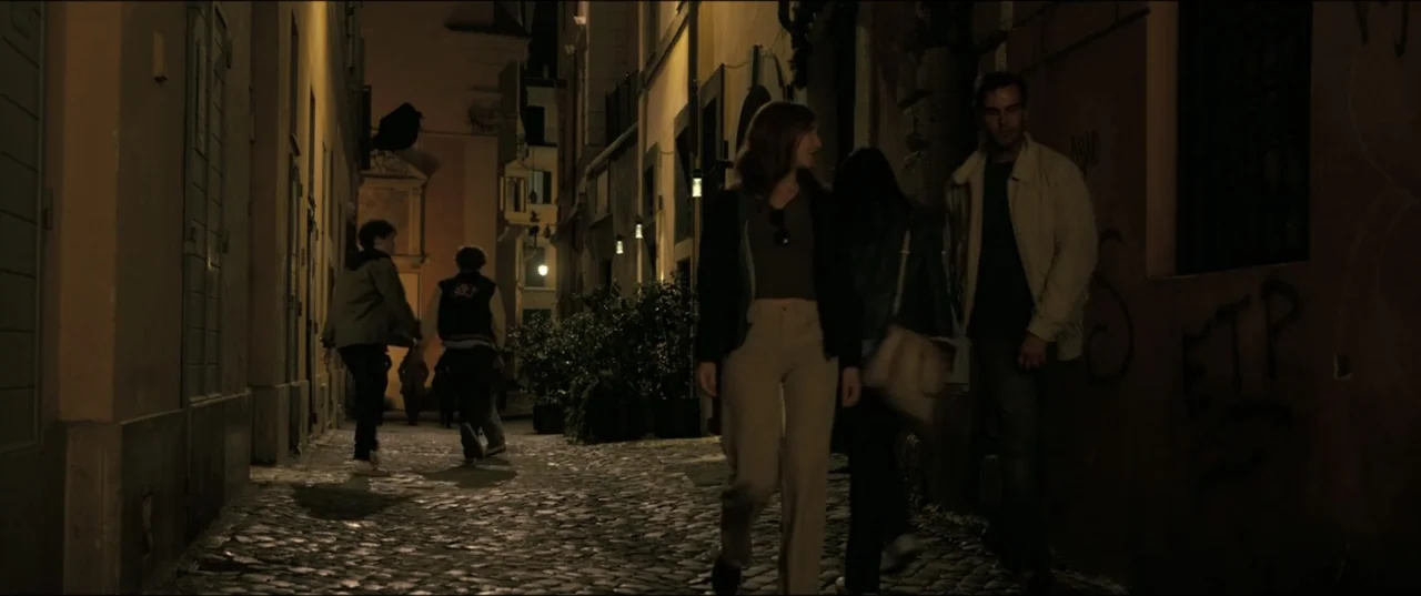 My Spy The Eternal City (2024) movie still: fullbody — A narrow cobblestone alleyway is flanked by tall buildings with dim warm artificial lighting.…