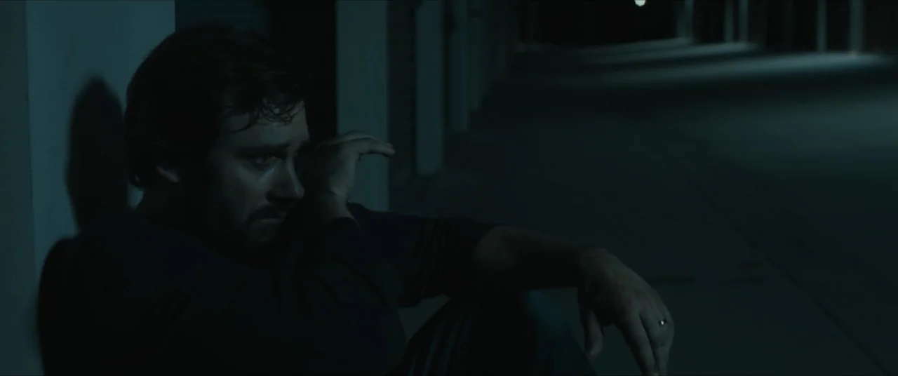 Possessions (2024) movie still: medium — A dark concrete parking garage at night with artificial light casting long shadows. A man sits on the floor…