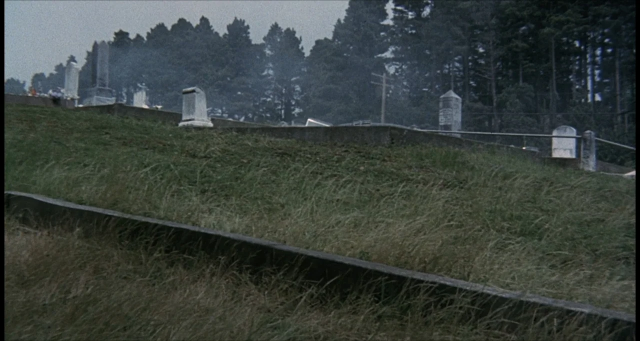 The Dunwich Horror (1970) movie still: establishing — An outdoor cemetery sits on a grassy hill under a cloudy sky. Multiple stone grave markers are…
