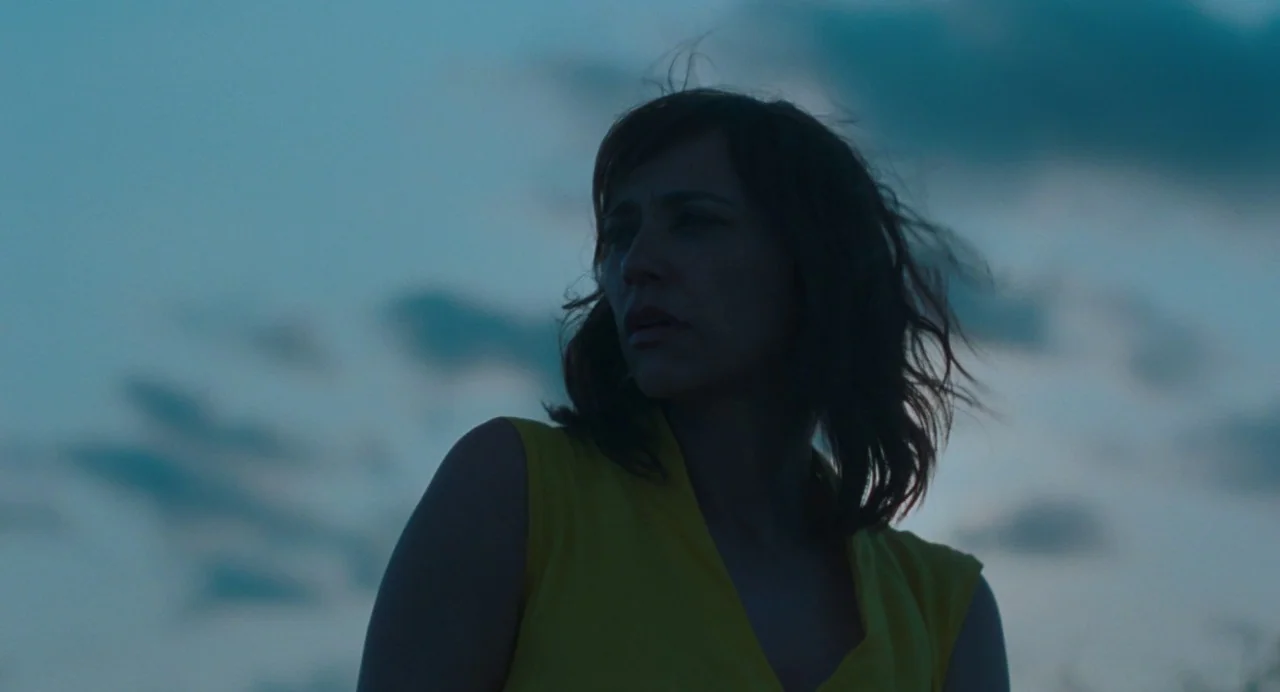 On The Rocks (2020) movie still: medium — An open landscape at dusk with a clouded blue sky. A woman looks off-camera while wearing a bright yellow…