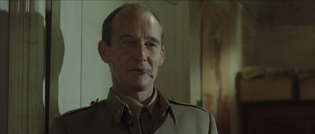 Legionario (2025) movie still: medium — A dimly lit room with plain walls features an indistinct blurred painting in the background. A man in a…