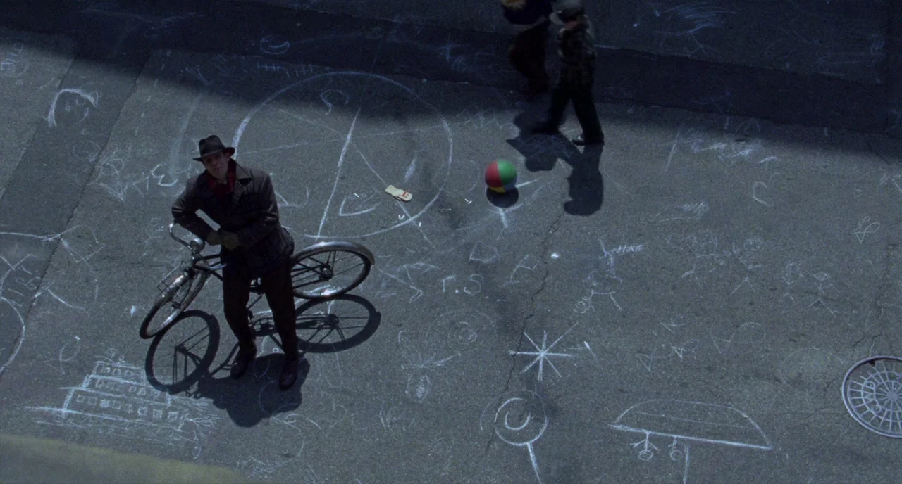 Carol (2015) movie still: wide — A paved asphalt surface covered in intricate white chalk drawings of shapes and symbols. A man in a fedora stands…