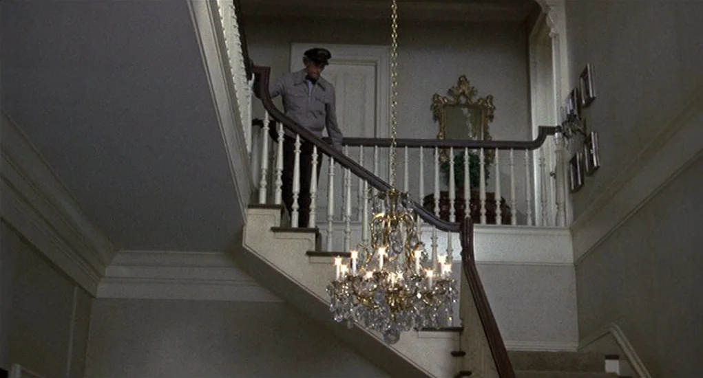 Family Plot (1976) movie still: wide — A grand residential staircase features a white railing and a prominent crystal chandelier hanging. An older…