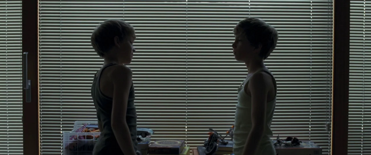 Goodnight Mommy (2014) movie still: medium — Two young boys stand facing each other against a window covered by horizontal blinds. Various toys are…