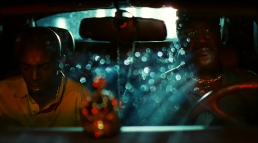 Clockers (1995) movie still: medium — Two men sit in the front seats of a car at night during rain. Bokeh lights scatter across the windshield as…