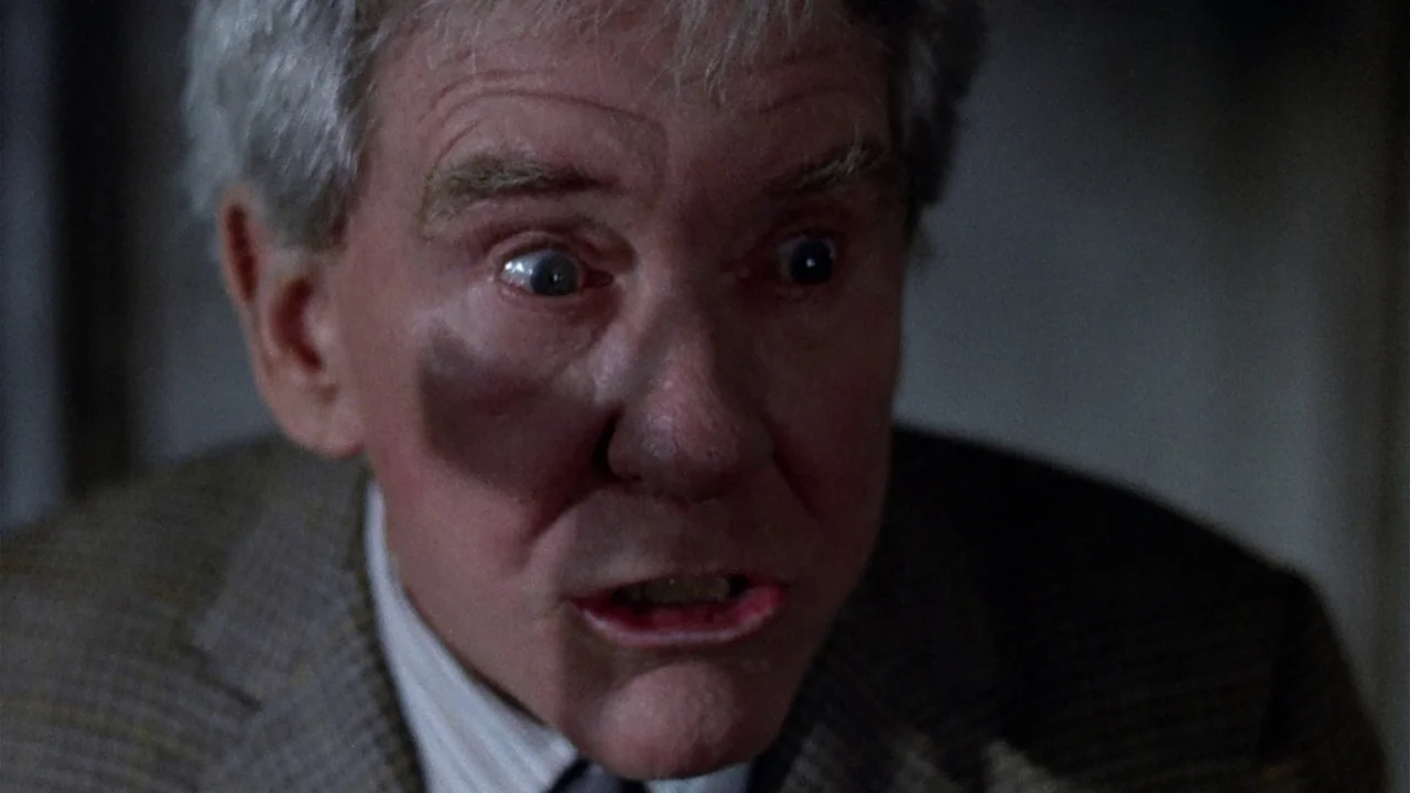 The Sentinel (1977) movie still: closeup — A close view of an elderly man with a bruised face in a dark room. The man stares forward with wide eyes…