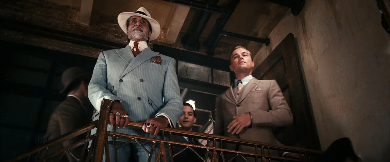 The Great Gatsby (2013) movie still: medium — A 1920s interior balcony with a metal railing and high ceilings overhead. Three men in formal suits…