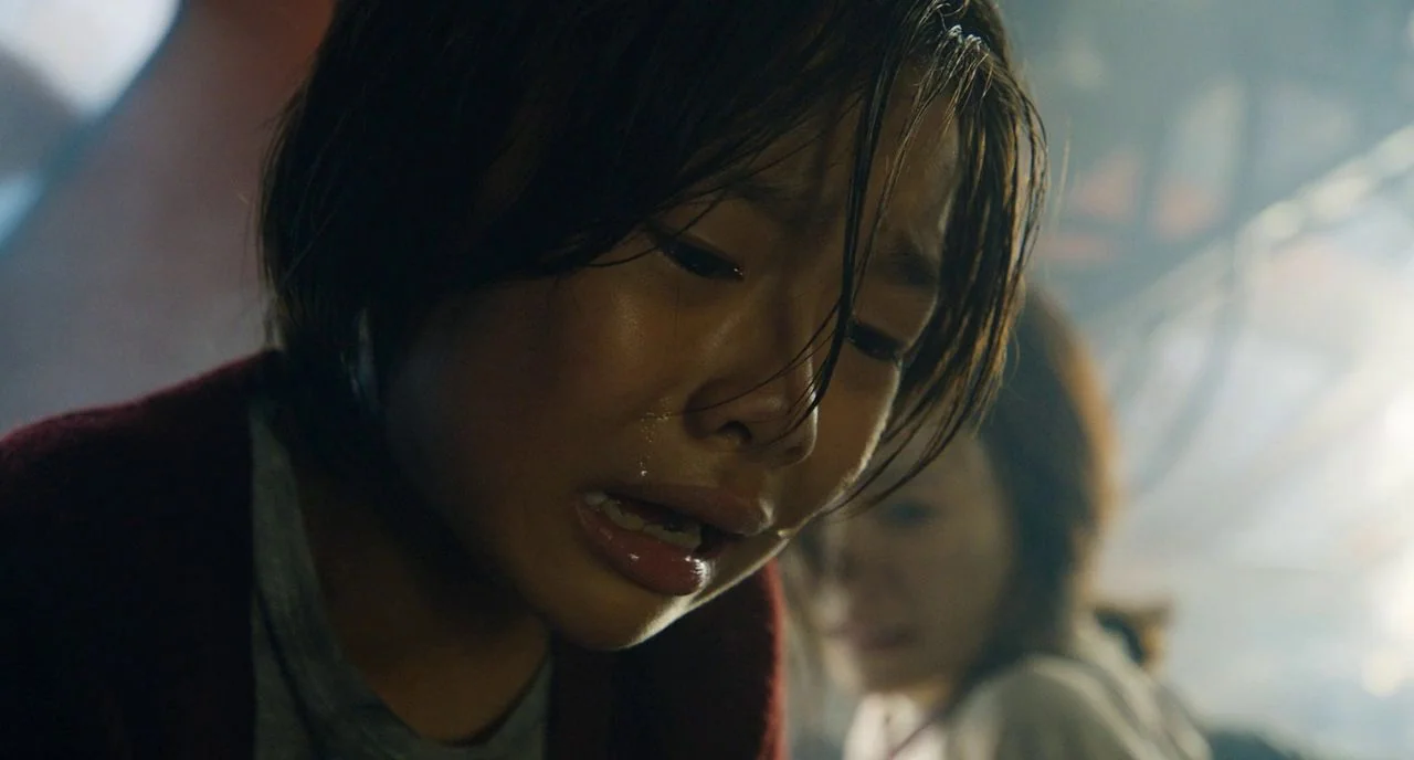 Train To Busan movie still: closeup — The interior of a vehicle is dark and filled with hazy smoke. A young child cries with their eyes closed and…