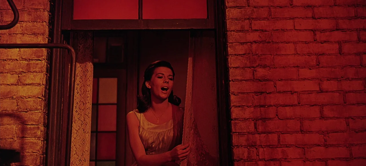 West Side Story (1961) movie still: medium — A dark brick building facade frames a woman standing at a window aperture. The woman gazes outward with…