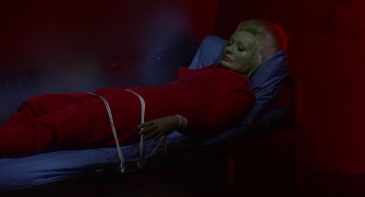 Queen of Blood (1966) movie still: medium — A dark, dimly lit room contains a narrow cot with blue bedding. A woman in a red jumpsuit lies bound to…