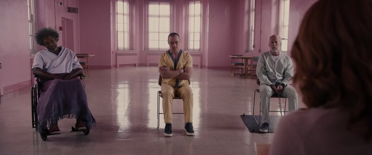 Glass (2019) movie still: wide — A spacious room with pale pink walls and large windows contains three men sitting. The men are positioned on chairs…