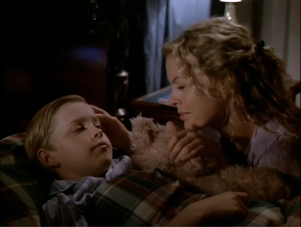 At the Midnight Hour (1995) movie still: medium — A dimly lit bedroom features a child lying in a bed with plaid covers. A woman leans over the bed…
