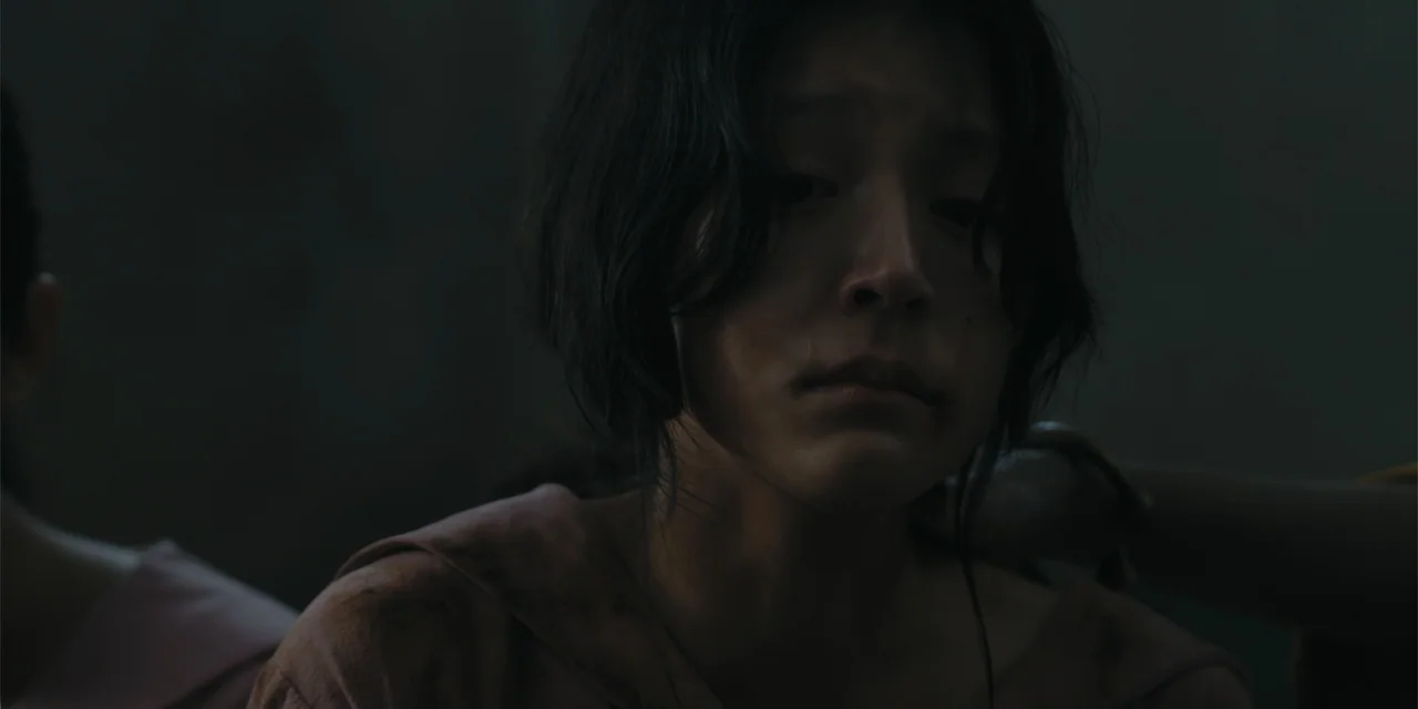 Host (2020) movie still: closeup — A dimly lit indoor space with blurred background surfaces. A young woman with dark hair sits quietly while an…