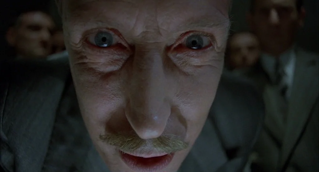 Brazil (1985) movie still: closeup — An elderly man peers closely into the camera in a darkened interior setting. Several other figures in suits…
