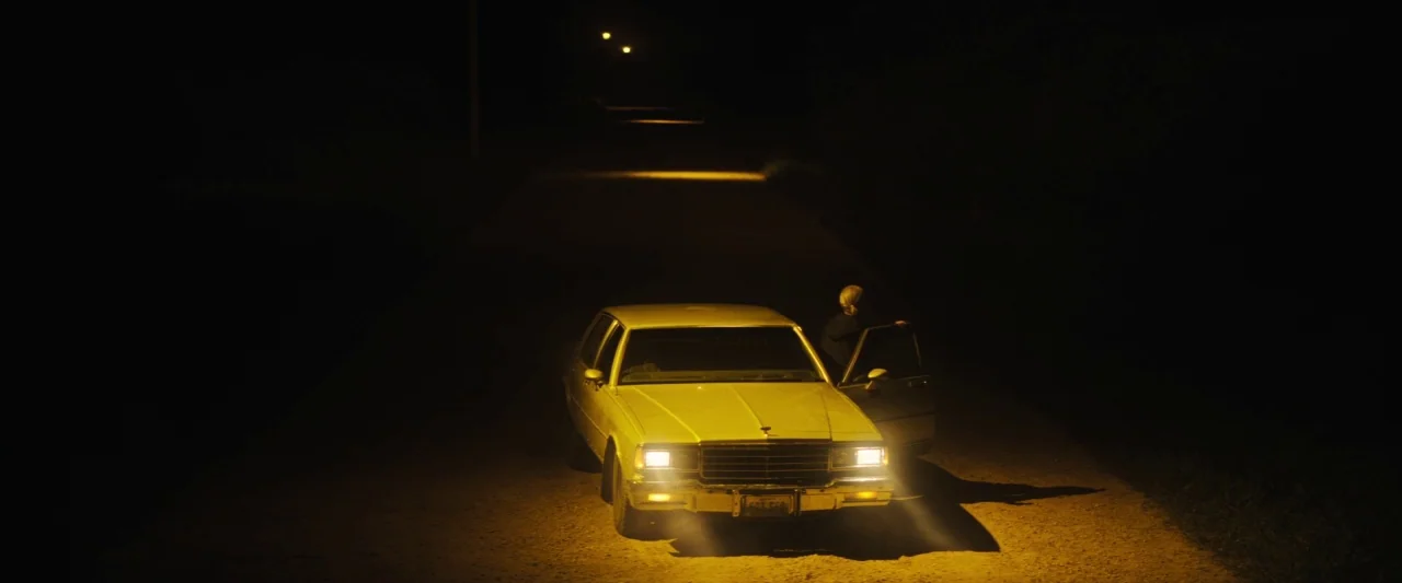 The Grudge (2020) movie still: wide — A vintage sedan sits stationary on a dark dirt road at night. The driver side door is open as the man exits…