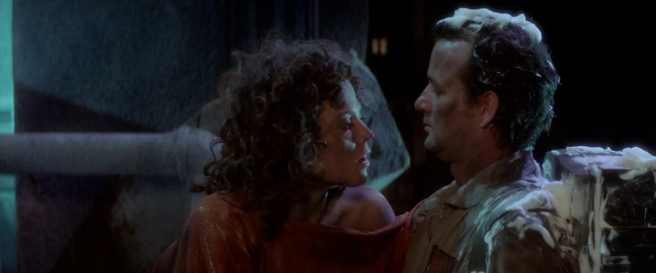 Ghostbusters (1984) movie still: medium — An interior space features dark walls and industrial pipes in the background. A woman and a man covered in…