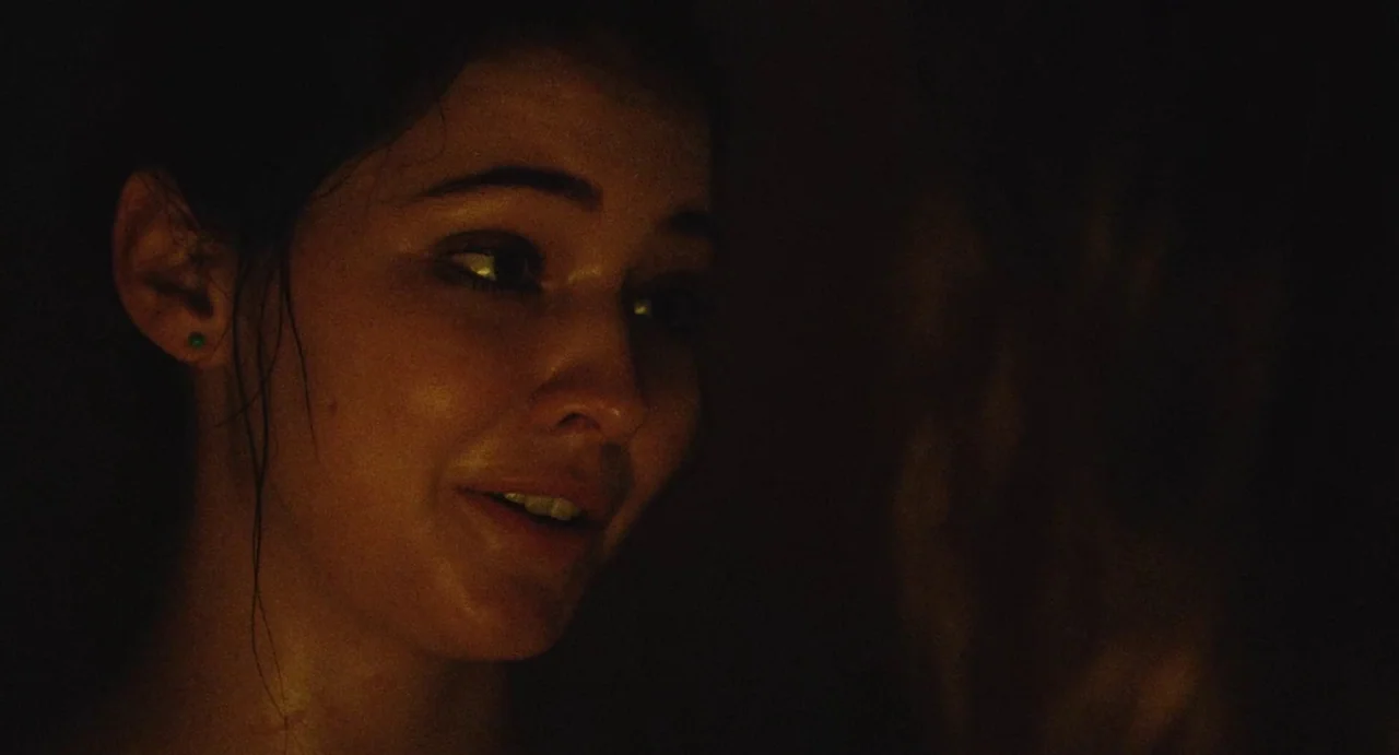 The Giant (2019) movie still: closeup — A dimly lit interior space with a subject captured in deep shadows. The woman turns her head slightly while…