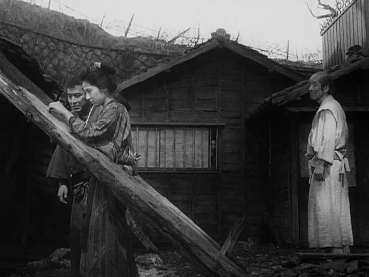 The Lower Depths (1957) movie still: fullbody — A rural setting features weathered wooden houses with barred windows and stone retaining walls. A…