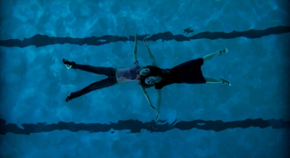 Water Lillies (2007) movie still: wide — A deep blue water surface marked by dark parallel floor lines of a pool. Two women float on their backs in…