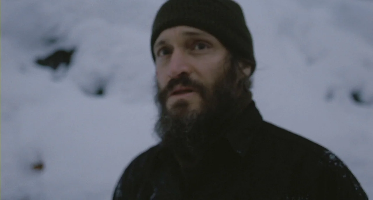 Essential Killing (2010) movie still: closeup — A man stands outdoors in a snowy environment with soft, flat lighting conditions. He looks toward…