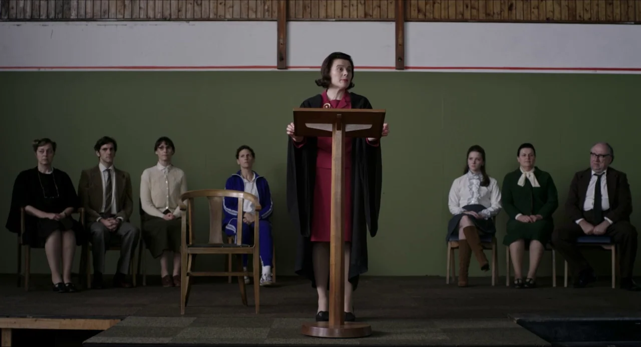 The Falling movie still: fullbody — A woman stands behind a wooden lectern on a school assembly hall stage. Seven adults sit in a row of chairs…