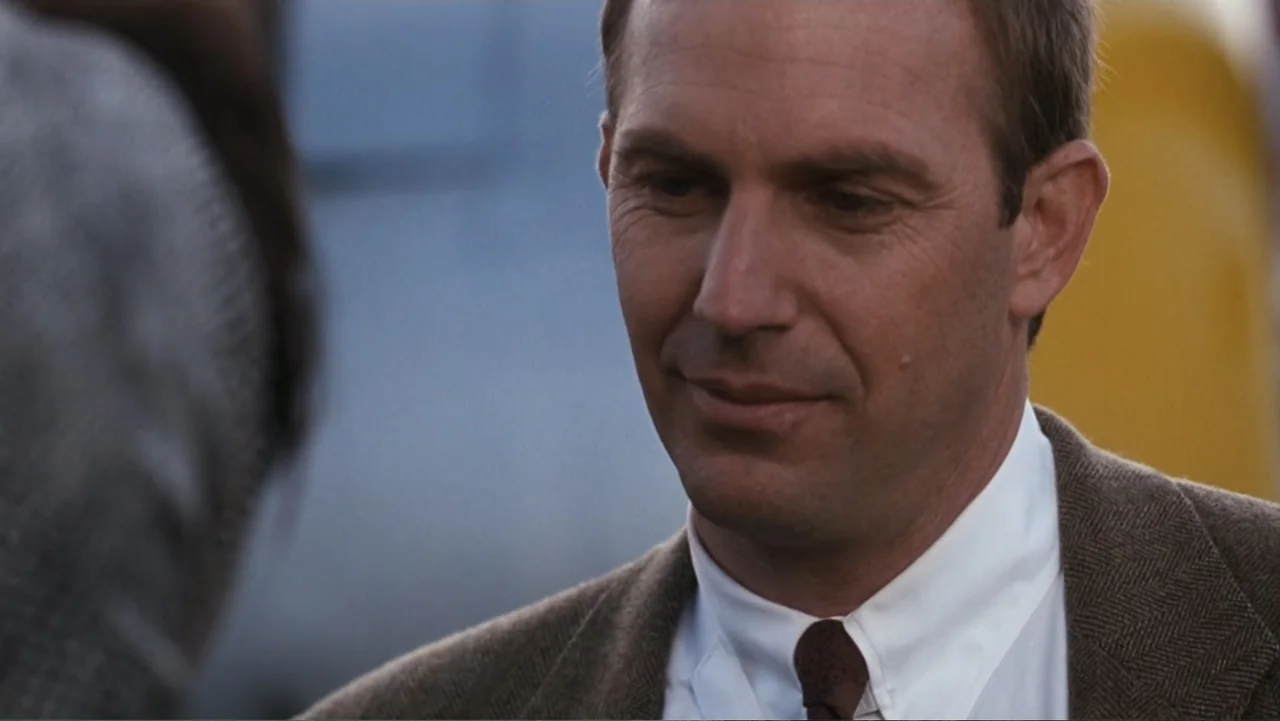 The Bodyguard (1992) movie still: closeup — A man in a brown tweed jacket stands at an outdoor airfield setting. The man turns his head slightly…