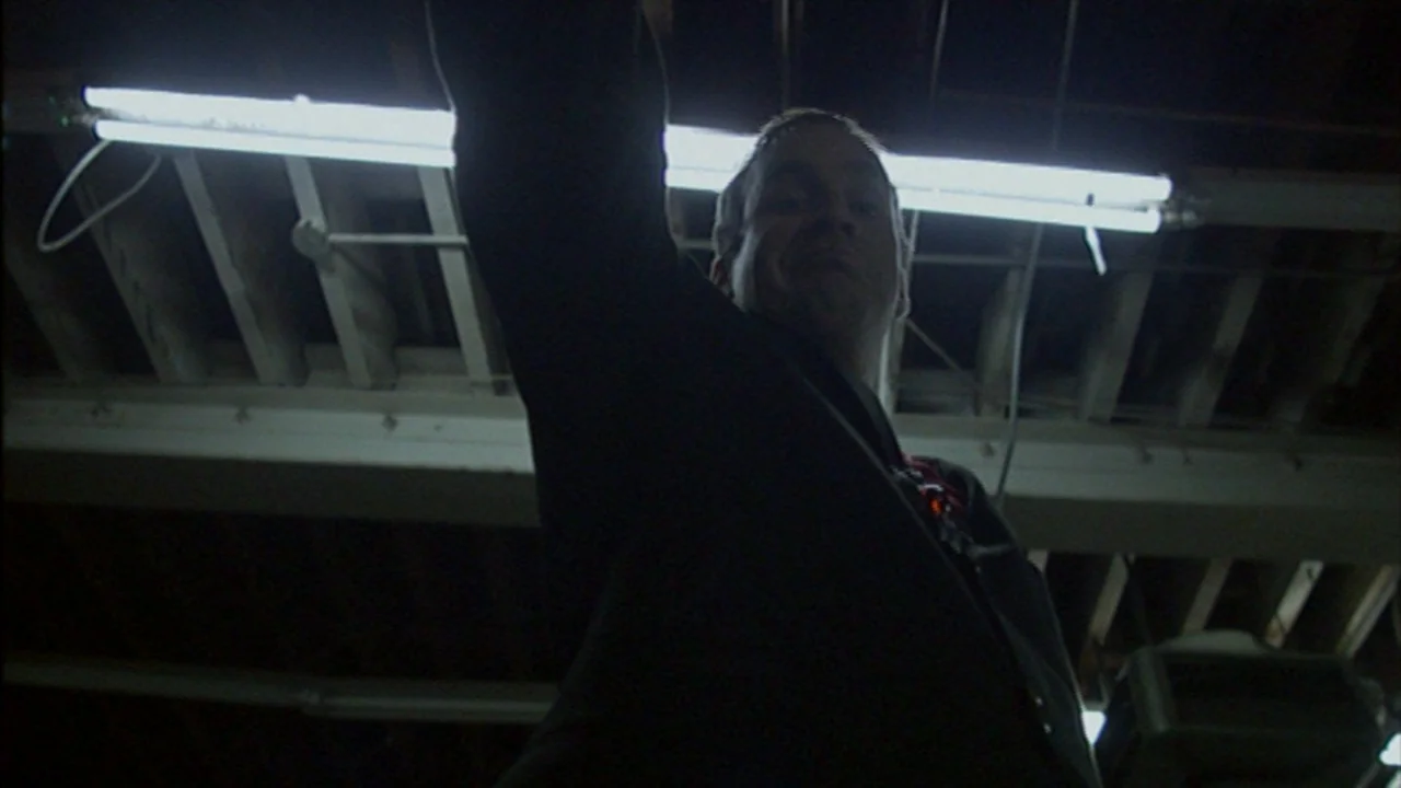 Murder Party movie still: medium — An unfinished basement with exposed wooden joists and harsh overhead lighting. A man stands below the camera…
