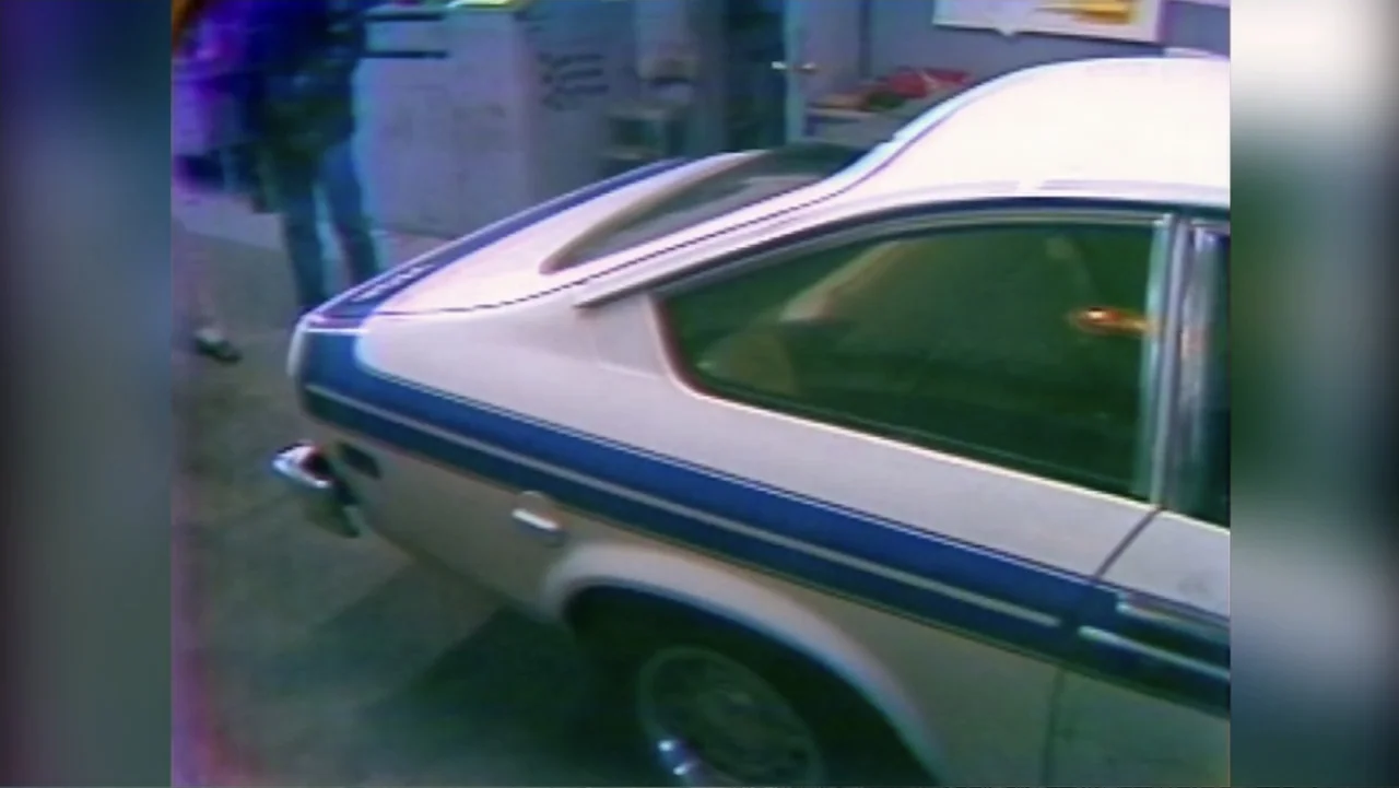 Murders at the Burger Joint (2022) movie still: medium — A vintage two-door sedan with blue and gold stripes sits on a gray surface. The rear end…