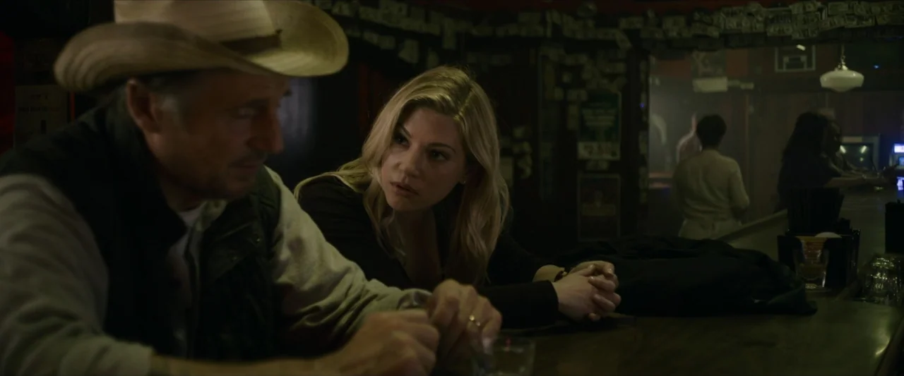 The Marksman (2021) movie still: medium — A dimly lit bar interior features wood paneling adorned with dollar bills on the walls. A man in a cowboy…