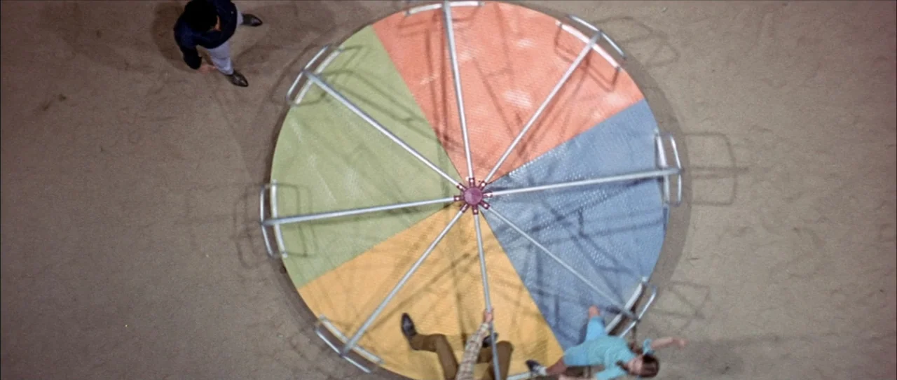 Clambake (1967) movie still: wide — A top-down view of a colorful circular carousel located in a sandy play area. Three people are positioned around…