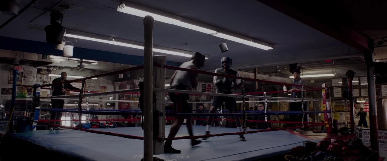 Creed (2015) movie still: wide — A basement boxing gym features a central ring surrounded by old posters and equipment. Two male boxers move across…