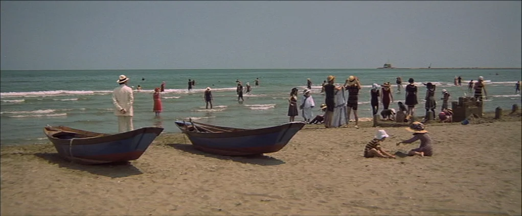 Death In Venice (1971) movie still: wide — A sunny 1920s beach scene features wooden boats resting on the sand. Numerous people in period swimwear…