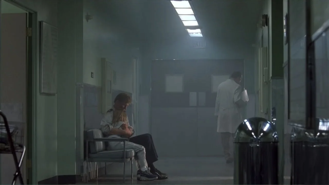 Crisis Point (2012) movie still: wide — A dimly lit hospital hallway features faded green walls and a distant exit door. A man sits on a metal…