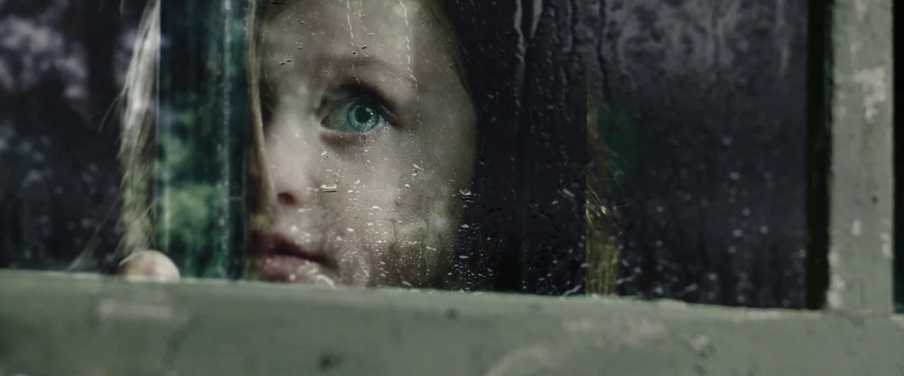 Wildling (2018) movie still: closeup — A child peers through a wet, dark window frame during a rainy night. A single wide eye is clearly visible…