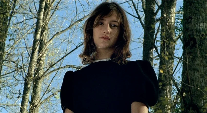 Bluebeard (2009) movie still: medium — A dense forest setting with tall trees and clear blue sky visible above. A young woman stands looking down…
