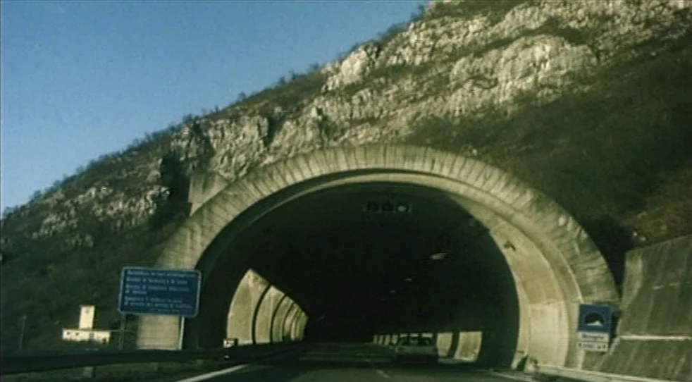 Scarlet Diva (2000) movie still: establishing — A concrete road tunnel bores through the base of a steep, rocky mountain slope. A lone car…