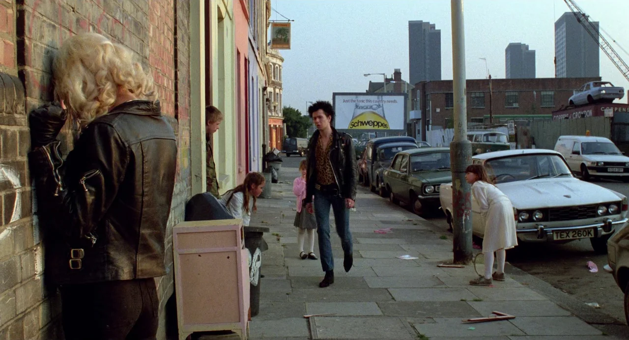 Sid and Nancy (1986) movie still: wide — A sidewalk in front of colorful buildings with parked cars and a distant billboard. A man with spiky hair…