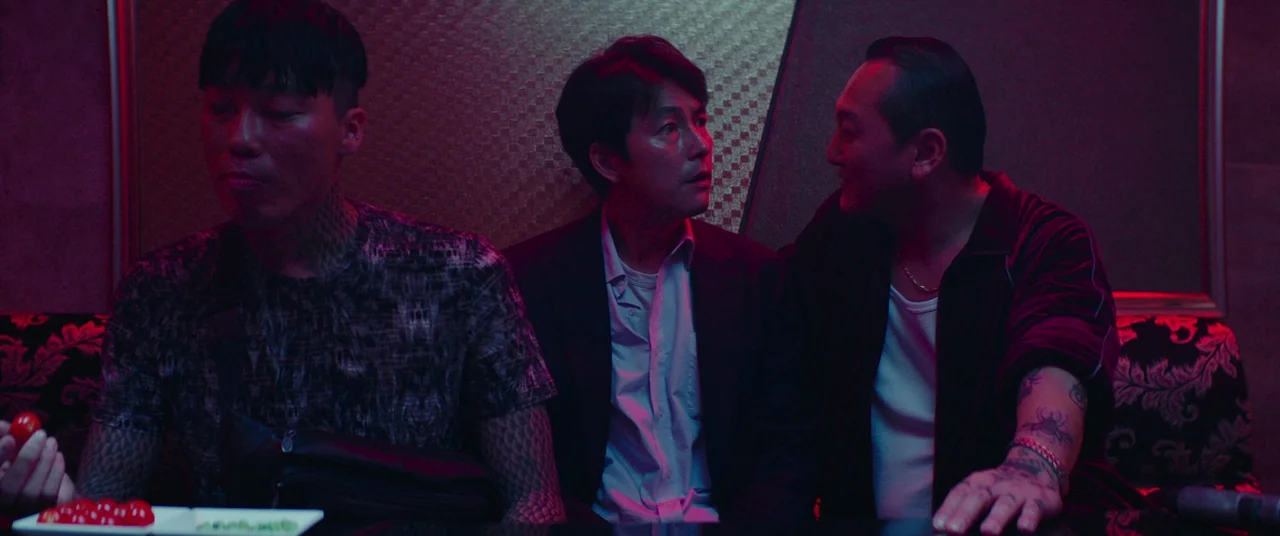 Beasts Clawing At Straws (2020) movie still: medium — A dark lounge booth with ornate patterned upholstery and dim ambient club lighting. Three men…