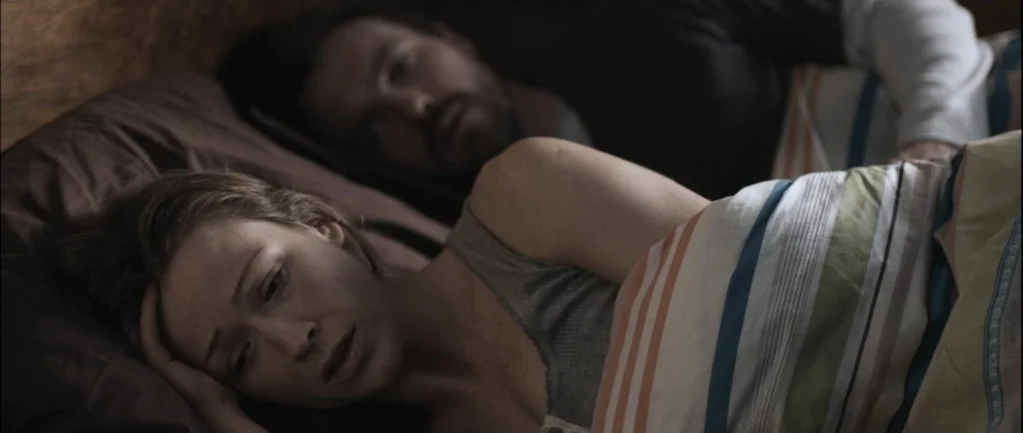 One Hundred Mornings (2009) movie still: medium — Two individuals lie side-by-side on a pillow in a darkened sleeping area. The woman is awake and…