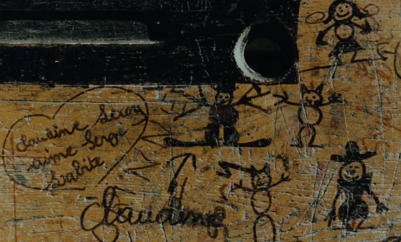 Graduate First (1978) movie still: closeup — A wooden desktop surface is covered in deep scratches and crude black ink drawings. Various stick…
