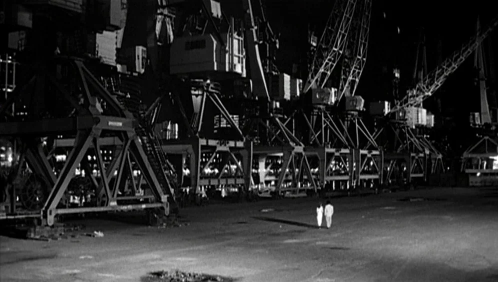The Tango Lesson (1997) movie still: wide — A vast industrial terminal at night features rows of massive metal shipping cranes. Two small figures…