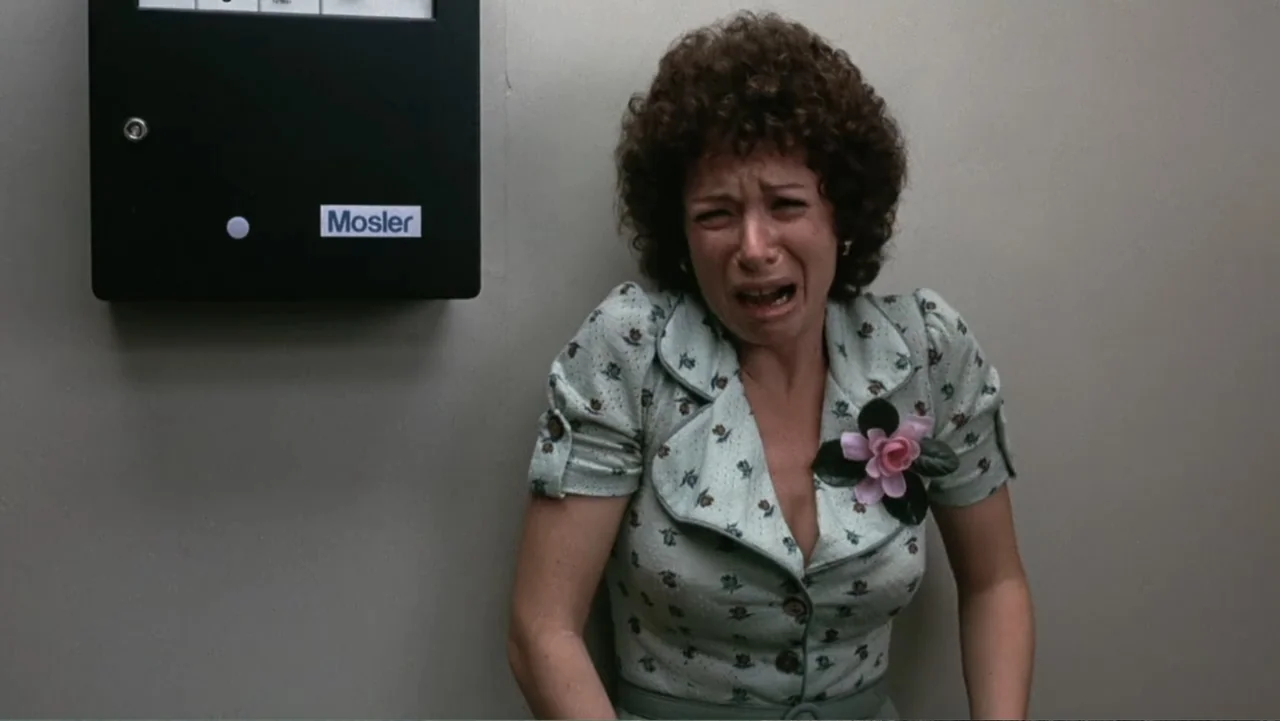 Dog Day Afternoon (1975) movie still: medium — A plain grey wall features a large black metal Mosler lock unit. A woman wearing a green patterned…
