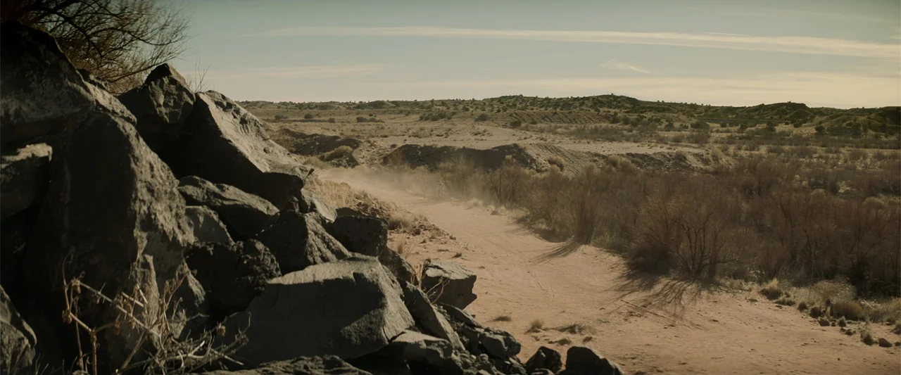Cry Macho (2021) movie still: wide — An arid desert landscape features a dirt path winding through rocks and brush. The sunlit terrain extends…