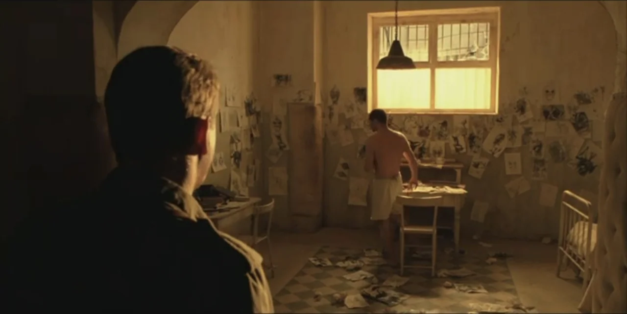 Exorcist: The Beginning (2004) movie still: medium — A sparsely furnished stone room contains a metal cot, desk, and drawings on walls. A man in a…