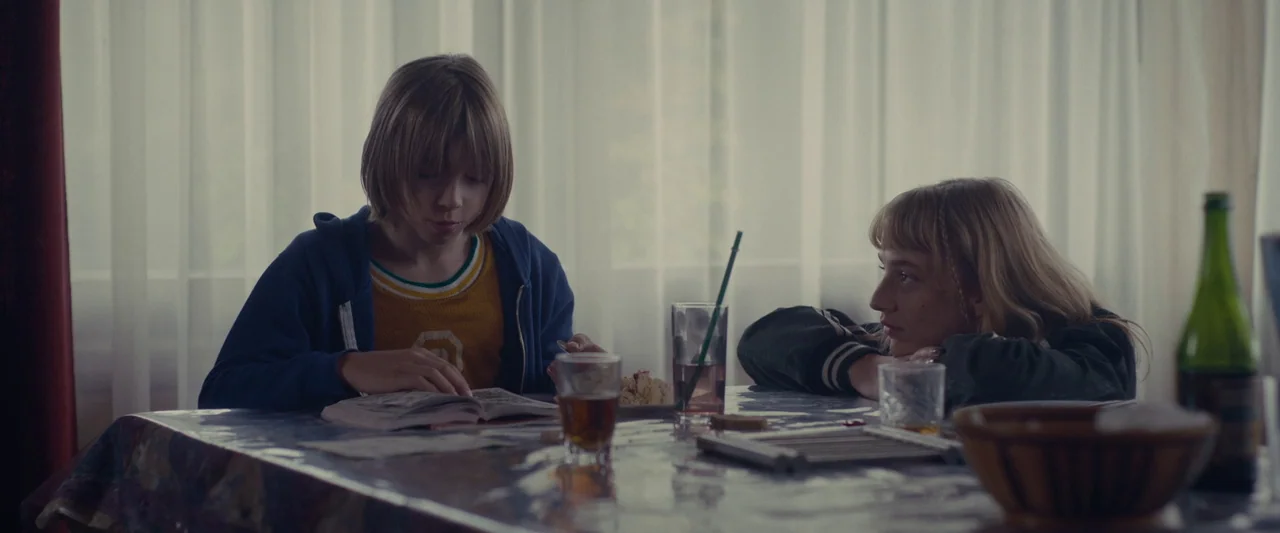 Meanwhile On Earth (2024) movie still: medium — A dining table with a plastic tablecloth is set with glassware and a bowl. A young boy reads a book…