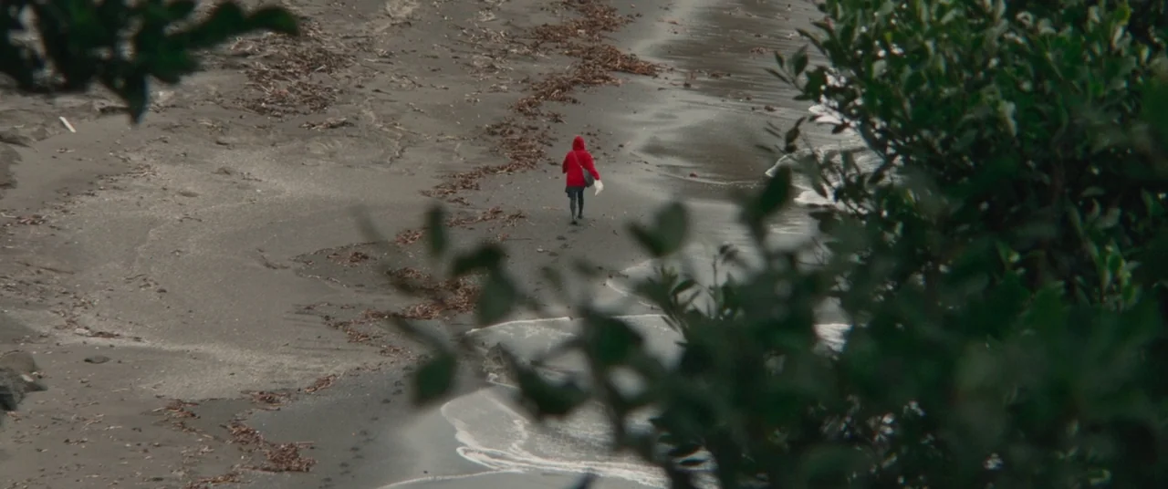 Kumiko the Treasure Hunter (2014) movie still: wide — A grey sand beach is littered with scattered driftwood and debris. A woman in a red hoodie…