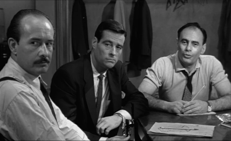 12 Angry Men (1957) movie still: medium — An interior space with three men seated around a wooden table in a room. The men look forward, one holding…