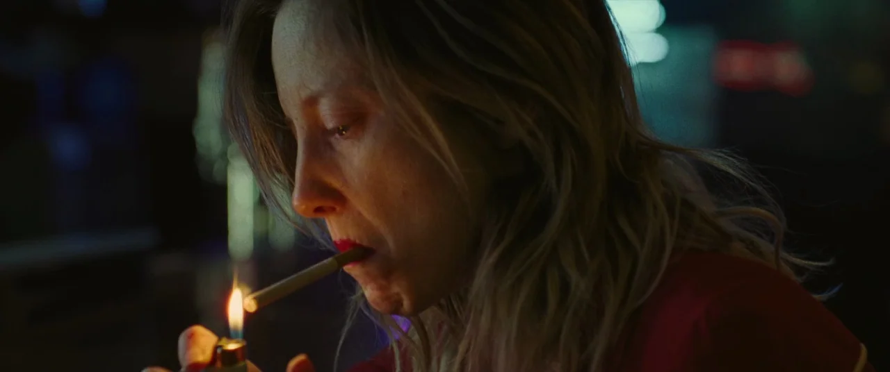 To Leslie (2022) movie still: closeup — A woman sits in a dark space lit by the flicker of a lighter. She holds a cigarette to her mouth while…